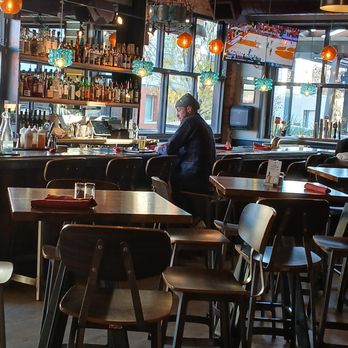 Acova - The Warmth of a bar surrounded by windows - A man wearing a dark jacket and cap is seated at a bar counter in a restaurant with large windows allowing natural light. The interior features colorful hanging lights, a well-stocked bar with bottles, and a TV screen displaying a sports game. The seating includes bar stools and tables with chairs, and the space has a modern, rustic decor with exposed beams and wooden accents. - New American Near Me - Denver, Colorado