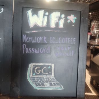 Ground Central - The chalkboard sign has a black frame and surface, with colorful chalk writing indicating Wi-Fi network and password details. It also includes a simple drawing of a laptop with the letters 'GC' on the screen. The sign is placed indoors, with a bright light source behind it, and part of a menu or display board visible in the background. - Coffee & Tea Near Me - Lynbrook, New York