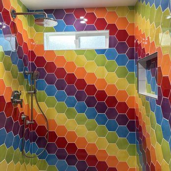 Method Remodeling - The new shower, freshly completed, before glass installation - A shower room with walls covered in vibrant, rainbow-colored hexagonal tiles arranged in a repeating pattern. The space includes a small horizontal window near the ceiling, allowing natural light. There are two showerheads: a fixed overhead rain shower and a handheld showerhead, both mounted on the wall. The floor has light-colored hexagonal tiles, and a recessed shelf is built into one wall for toiletries. - General Contractors Near Me - Burlingame, California