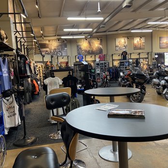 Willamette Valley Harley-Davidson - The image depicts the interior of Willamette Valley Harley-Davidson, a motorcycle dealership. It includes motorcycles displayed on the floor, merchandise such as clothing and accessories on shelves and racks, and seating areas with tables and chairs. The space is well-lit with overhead lighting, and promotional posters and photographs are visible on the walls. The dealership serves the Willamette Valley region, including Eugene and Portland. - Motorcycle Dealers Near Me - Eugene, Oregon