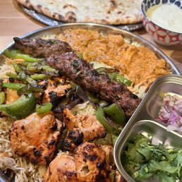 Chicken Tikka Masala - The image shows a large metal platter filled with Indian cuisine, including grilled chicken, lamb kebabs, rice, and sautéed green peppers and eggplant. A piece of naan bread is placed on a separate tray in the background. A bowl of yogurt-based sauce and a small container with chopped onions and cilantro are also visible. The meal is served on a wooden table, typical of a restaurant setting.