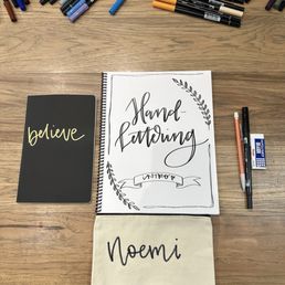 The materials she provides. - A collection of markers, pens, and a notebook with hand-lettered text on a wooden table, used for hand lettering practice.