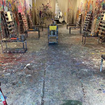 The Splatter Studio - The image shows an art studio with walls covered in multicolored paint splatters. Metal chairs with paint on their surfaces are arranged along the sides, each with paint rollers placed on the seats. The floor is heavily stained with various colors of dried paint, and there are paint cans and tools on small tables. Curtains at the back of the room partially obscure the view of additional art supplies or workspace beyond. The setting appears to be a space for abstract or splatter painting activities. - Team Building Activities Near Me - Atlanta, Georgia
