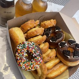 Some delicious donuts, Oreo cookie, old fashioned, sprinkles, maple and chocolate bars. Apple juice and coffee. - The image shows a cardboard takeout box filled with assorted donuts and fried pastries, including a donut with colorful sprinkles, chocolate-covered donuts with Oreo pieces, and fried items. Three bottled beverages are placed behind the box, and the setting appears to be on a table with a patterned placemat and a tablecloth. The scene suggests a casual snack or dessert arrangement.