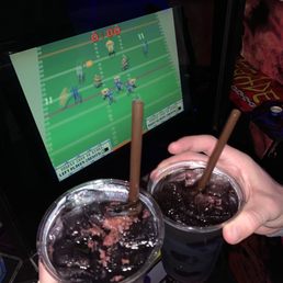 The Scoundrel - Two individuals hold cups of dark soda with straws in front of a pixelated arcade game screen at Bar Botics, a retro-themed arcade bar.