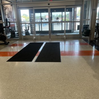 DICK'S Sporting Goods - The photo depicts the entrance of a retail store with large glass sliding doors, metal frames, and a view of the parking lot outside. Two black mats are placed on the beige and orange-tiled floor just inside the entrance. The parking lot has several cars and trees in the background. The interior includes some gym equipment on the left and a shopping cart on the right. The lighting is natural, coming through the glass doors. - Sports Wear Near Me - Massapequa, New York