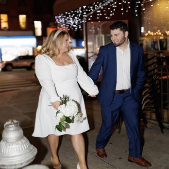 Sotto Voce - A woman in a white dress holding a bouquet of white flowers and a man in a blue suit walk hand-in-hand on a city sidewalk at night. The woman has blonde hair and is wearing white heels, while the man has dark hair and is wearing brown shoes. They are smiling at each other. The background features a restaurant with a red awning and string lights, with some parked cars and illuminated signs visible. - Italian Near Me - Brooklyn, New York