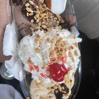 Scoops - Brownie Sundae - A clear plastic container contains several scoops of ice cream in different flavors, topped with chopped nuts, chocolate syrup, and a cherry. A white spoon is inserted into one of the scoops, and a napkin lines the container. The image captures the variety of toppings and the presentation of the ice cream dessert. - Ice Cream & Frozen Yogurt Near Me - Smithfield, Rhode Island