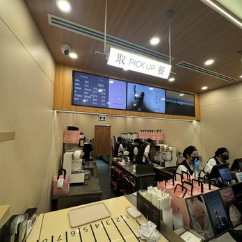 Molly Tea - interior - The image shows a food pickup counter inside a modern establishment, with staff members wearing uniforms and masks working behind the counter. Above, a sign with Chinese characters and the words 'PICK UP' indicates the area for collecting orders. Digital screens display flight information or queue numbers, suggesting a themed or airport-like setting. The foreground features a numbered counter surface, and various equipment and supplies are visible on the counters. The ceiling has recessed lighting and wooden paneling, creating a clean and organized environment. - Bubble Tea Near Me - Markham, Ontario