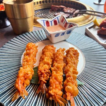 Y Space BBQ - Fried shrimp - too much batter, skippable - Four fried shrimp skewers with golden, crispy batter are placed on a decorative black-and-white striped plate, with a small bowl of dipping sauce nearby. The background shows a grill with meat cooking, along with other food items and utensils, indicating a barbecue restaurant setting. The image captures a close-up of the fried seafood dish and part of the dining environment. - Barbeque Near Me - Flushing, New York