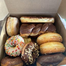 A box contains various donuts such as glazed, chocolate-covered, sprinkled, and filled donuts, arranged in a cardboard container.