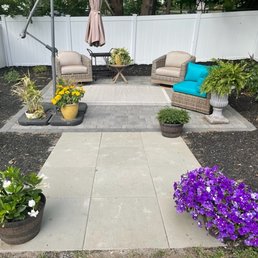 A backyard patio features two beige armchairs, a wicker sofa with a blue cushion, and a small table with potted plants, surrounded by flower beds and colorful blooms.