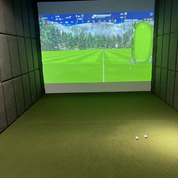 Golfinity - An indoor golf simulation room features a large screen showing a golf course landscape, with two golf balls on the green carpeted floor. - Golf Lessons Near Me - Austin, Texas