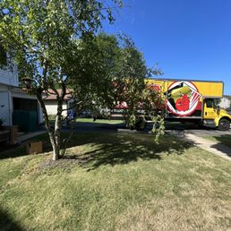 Loading up at our old house - A yellow moving truck with a graphic logo is parked in a residential driveway next to a house. A tree with green leaves partially obscures the truck, and the lawn in the foreground has patches of sunlight and shadow. The scene is set on a clear day with a bright blue sky.