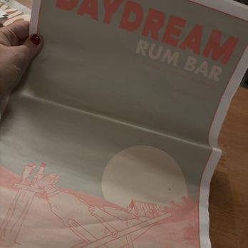 Daydream - This is the menu! Super fun! - The image depicts a menu for Daydream Rum Bar, with a beach-themed illustration. The illustration shows a person reclining on a lounge chair, holding a drink, with a large sun or moon in the background. The menu is held by a hand with red nail polish, and the setting appears to be indoors, possibly at a bar or restaurant. - Cocktail Bars Near Me - Albuquerque, New Mexico