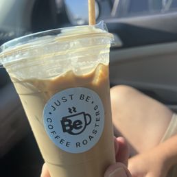 carmel latte - A caramel latte in a clear plastic cup with a lid and a straw, displaying the 'Just Be Coffee Roasters' logo. The drink appears to be creamy and topped with a layer of foam. The cup is held by a person's hand inside a vehicle, with the car's interior and a parked vehicle visible in the background.