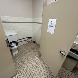 Del Lanes - An accessible toilet stall, with a sign saying out of order. There is a hole in the wall where some plumbing is missing.