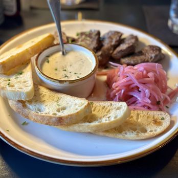 Nyla's Restaurant - Beef tips fondue - A round ceramic plate contains beef tips, sliced and cooked, accompanied by several slices of toasted bread, a small bowl of creamy herb sauce, and a portion of pink pickled onions. The dish is served at Nyla's Restaurant in Westfield, IN, which offers American cuisine in a rustic setting. The background shows a dark table surface and some glassware. - Cocktail Bars Near Me - Westfield, Indiana
