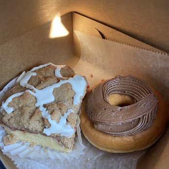 DeLuise Bakery - two doughnuts in a box - A cardboard box holds a crumb cake with icing and a chocolate frosted donut, placed on parchment paper. - Bakeries Near Me - Cranston, Rhode Island