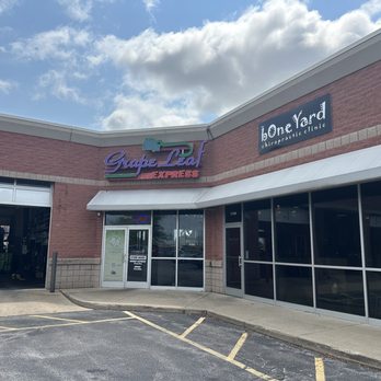 Grape Leaf Express - Exterior - The photo depicts the exterior of a strip mall featuring two businesses: Grape Leaf Express, a restaurant offering Lebanese, Greek, and American cuisine, and Bone Yard, a chiropractic clinic. The storefronts have large glass windows and doors, with signage above each entrance. The parking lot in front has marked parking spaces, and the sky above is partly cloudy. - Mediterranean Near Me - Toledo, Ohio