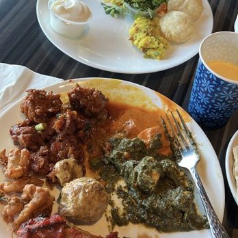 India 101 - A plate of Indian food includes multiple curries, rice, and bread, with a fork placed on the plate. In the background, there is a side dish with rice, chutneys, and salad, along with a cup of orange-colored drink and a blue patterned cup. The setting appears to be a restaurant with a dark wooden table. - Indian Near Me - Irving, Texas