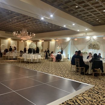 AGS Event Creations - The image shows a spacious banquet hall with round tables set for a formal event, decorated with white tablecloths and floral centerpieces. Elegant drapery and large floral arrangements adorn the background, with a dance floor in the foreground. The ceiling has a wooden pattern, and chandeliers provide lighting. Guests are seated and standing, engaging in conversations, indicating a social gathering such as a wedding reception or celebration. - Wedding Planning Near Me - Mundelein, Illinois