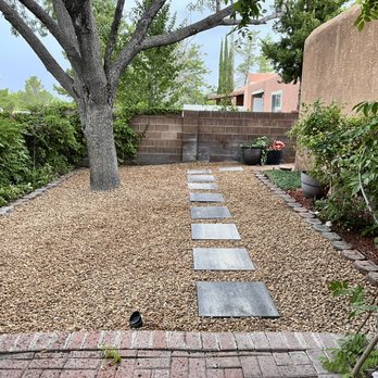AMG Landscape - Backyard with walking steps. - A landscaped backyard features gravel ground, a central tree, stepping stones, and potted plants along the fence wall. - Irrigation Near Me