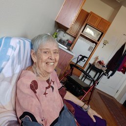 We found her smile - A smiling elderly woman with short gray hair, wearing a patterned blouse and purple pants, is seated in a wheelchair in a home kitchen. The background includes wooden kitchen cabinets, a microwave, a sink, and various household items. The woman appears relaxed and happy, with her arm resting on the wheelchair armrest.