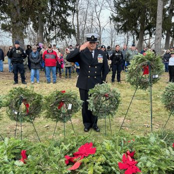 Sheeba Restaurant - A military officer dressed in a formal uniform with medals salutes during a wreath-laying ceremony. Several wreaths decorated with red ribbons are arranged in front of him on stands. A group of people, including adults and children, stand in the background, observing the event in an outdoor area with trees and overcast sky. - Middle Eastern Near Me - Dearborn, Michigan