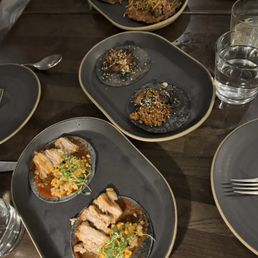 Carne Asada Tacos - The image shows a dining table with several dark-colored plates containing Latin-inspired dishes. The main plate features sliced meat with a vegetable garnish, while other plates hold small, garnished bites and appetizers. There are glasses of water and utensils visible, indicating a meal setting in a restaurant with modern tableware.