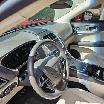 Masterwash Mobile Auto Detial - A detailed view of a vehicle's interior, featuring a digital dashboard, steering wheel with control buttons, center console with cup holders, and front passenger seat. Sunlight enters through the windows, highlighting the beige and black color scheme and modern design elements. - Auto Detailing Near Me