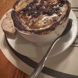French Onion Soup - A bowl of French Onion soup features a caramelized, slightly burnt cheese crust on top, with visible bubbling and charring. The soup is served in a white bowl placed on a plate, with a metal spoon resting beside it. A piece of bread is positioned on the plate next to the bowl, and the setting appears to be in a restaurant with a wooden table surface.