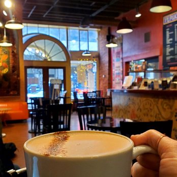 Perugino - chai latte - A person holds a white mug of chai latte inside a coffee shop with wooden tables and chairs, warm lighting fixtures, and large windows with decorative lighting outside. The interior features brick walls and artwork, creating a cozy atmosphere. - Cafes Near Me - Eugene, Oregon