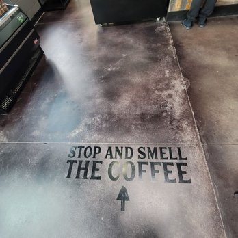 Vibrant Coffeehouse + Kitchen - There are little touches EVERYWHERE - The image shows a concrete floor inside a coffee shop with a decal that reads 'STOP AND SMELL THE COFFEE' in bold black and green letters. An arrow beneath the text points forward, guiding customers to the coffee area. The background includes a display case with baked goods, a person standing near the counter, and some equipment and shelving. The decal is positioned near the bottom center of the image, with the person's feet visible at the top right. - Coffee & Tea Near Me - Moline, Illinois