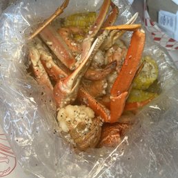 Pick 3 Seafood Boiler - Crab legs and pieces of corn on the cob are inside a clear plastic bag, which is placed in a takeout container. The seafood appears seasoned with spices and garlic. The background shows a paper tray and utensils, indicating a takeout or casual dining setting.