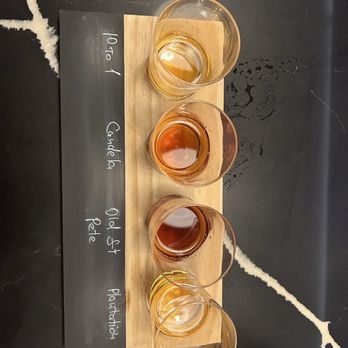 Rum Room - A set of five whiskey glasses is placed on a rectangular wooden board, each containing varying amounts of whiskey with different colors. The board has handwritten labels in white ink, indicating the whiskey types and aging periods, such as '10 to 1', 'Cask 6', 'Old St. Pete', and 'Plantation'. The background is a dark, textured surface with some white streaks and marks. - Cocktail Bars Near Me - Miami Beach, Florida