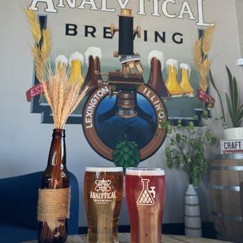 Analytical Brewing - Two glasses of craft beer, one light and one darker, are placed on a wooden table at Analytical Brewing. A decorative brown bottle wrapped with twine holds wheat stalks. The background features a mural with brewing equipment, hops, and the business name, along with potted plants and a barrel. The setting appears to be indoors with natural light illuminating the scene. - Beer Bar Near Me - Lexington, Illinois