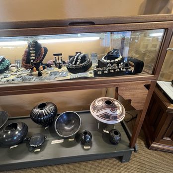 Taos Blue - Jewelry including necklaces, earrings, and bracelets are arranged inside a glass display case, with some pieces on black velvet busts and stands. Below, on a lower shelf, there are black and patterned pottery vases and bowls. A sign labeled 'Suzanne Betz' is visible above the display, indicating the artist or collection. The setting appears to be an art gallery or boutique specializing in handcrafted jewelry and ceramics. - Art Galleries Near Me - Taos, New Mexico