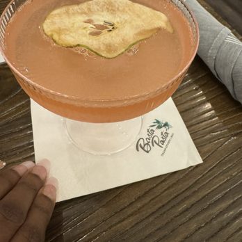 Basta Pasta - Fallston - Pearing is caring - A pink-colored cocktail with a slice of apple floating on top, served in a coupe glass on a wooden table at Basta Pasta in Fallston, MD. - Italian Near Me - Fallston, Maryland