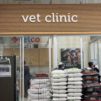 Petco - Vet Clinic's vaccines are offered on Sunday's! - The image shows the exterior of a vet clinic with a wooden slatted sign displaying 'vet clinic'. In front of the entrance, there are stacks of pet food bags, including brands like Wellness and others. The glass doors reveal the interior, where staff and a person wearing a mask are visible. A large poster of a person with a dog is also seen inside. The setting appears to be inside a shopping center or pet store area. - Pet Stores Near Me - Bakersfield, California