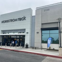 The photo depicts the exterior of a Nordstrom Rack store located in Oceanside, CA. The store's sign is prominently displayed above the entrance. There are a few people near the entrance, some sitting and some standing, with shopping carts visible. The building has a modern design with large windows and a clean facade. A parking lot with a vehicle and a blue inflatable object is also visible in the foreground. The overall scene is overcast with cloudy skies.