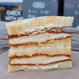 Spicy Truffle Chicken - A chicken katsu sandwich with three layers of bread, shredded cabbage, and sauce, served on a napkin outdoors.
