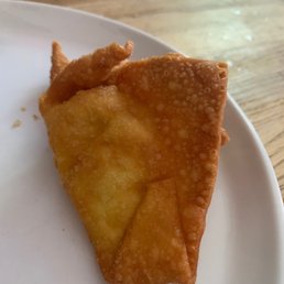 a piece of fried food on a plate - A fried wonton with a crispy, golden-brown exterior is placed on a white plate. The background shows a wooden table surface, indicating a casual dining setting. The wonton appears to be freshly cooked, with a textured surface and slightly bubbled edges.