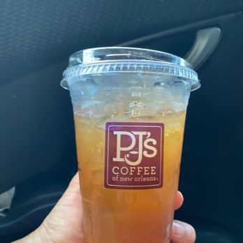PJ's Coffee - a hand holding a cup of iced coffee - A transparent plastic cup filled with iced tea from PJ's Coffee of New Orleans is shown, with ice cubes visible inside. The cup has a clear plastic lid and a purple logo with the text 'PJ's Coffee of New Orleans.' The background includes the interior of a vehicle, with a textured black surface and a door handle visible. The person holding the cup has a light skin tone and is gripping it with their fingers. - Coffee & Tea Near Me - Covington, Louisiana