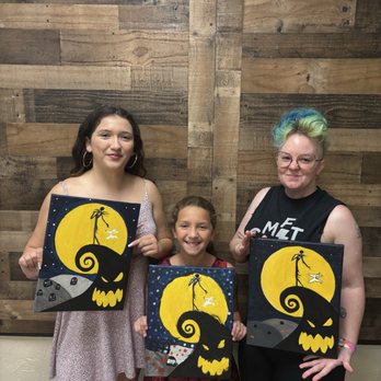 Paint & Sip Studio Temecula - Three women stand against a wooden wall, each holding a painting of a Halloween scene with a yellow moon, a witch on a broomstick, and a pumpkin face. - Paint & Sip Near Me - Temecula, California