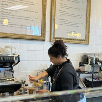 Jasmine Jo - A woman with dark, curly hair tied up in a bun is working behind the counter of a coffee shop, preparing a drink with lemon slices in a glass. The background features a white tiled wall with two framed menu boards listing coffee and food options. The counter has various coffee-making equipment, jars, and utensils. The scene captures a typical barista preparing beverages in a modern coffee shop setting. - Coffee & Tea Near Me - Tempe, Arizona