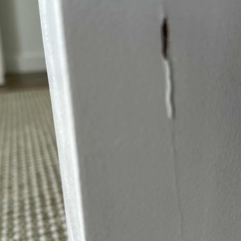 Skyroad Moving - Guest bed - The image depicts a gray painted wall with a vertical crack running down its surface and a small, dark hole nearby. The wall appears to be indoors, with a textured carpeted floor and a partial view of a door frame or furniture on the left side. The focus is on the damaged area of the wall, with the background slightly blurred. - Movers Near Me - Miami, Florida