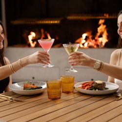 Gala Restaurant & Muse Bar - Two women toast with cocktails over dinner near a warm, glowing fireplace.