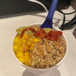 Matcha Goddess Smoothie Bowl with mangos, goji berries and oatmeal - A smoothie bowl in a white cup contains toppings such as diced mango, goji berries, and oatmeal. The bowl is placed on a white surface with visible cords and a black background. A blue spoon is inserted into the bowl, and the image captures the colorful toppings and the texture of the ingredients.