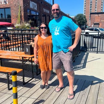 Notch Brewing - A man and woman pose outdoors on a wooden deck in front of a brick building with a sign for a tap room, under a clear blue sky. - Breweries Near Me - Salem, Massachusetts